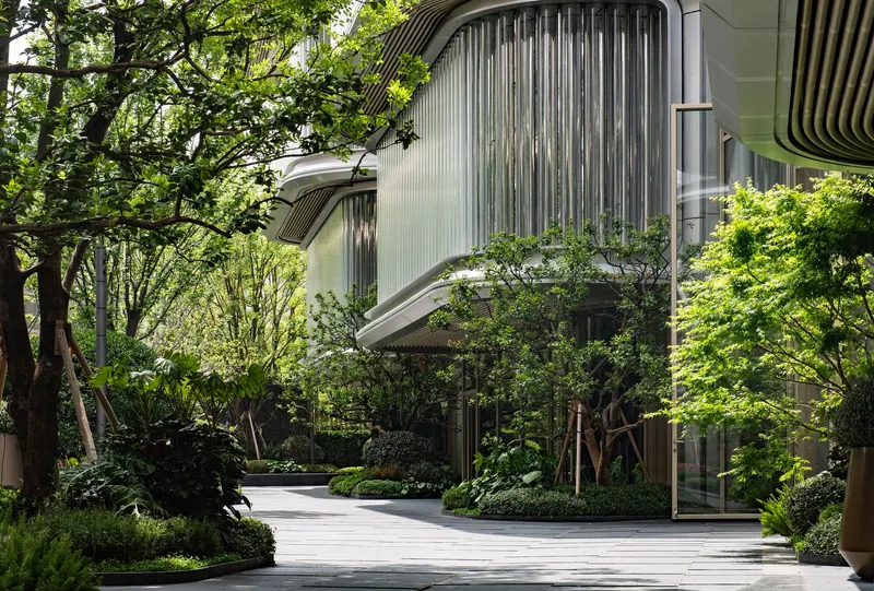 Curved facade with vertical louvers viewed from a landscaped courtyard path surrounded by trees in spring