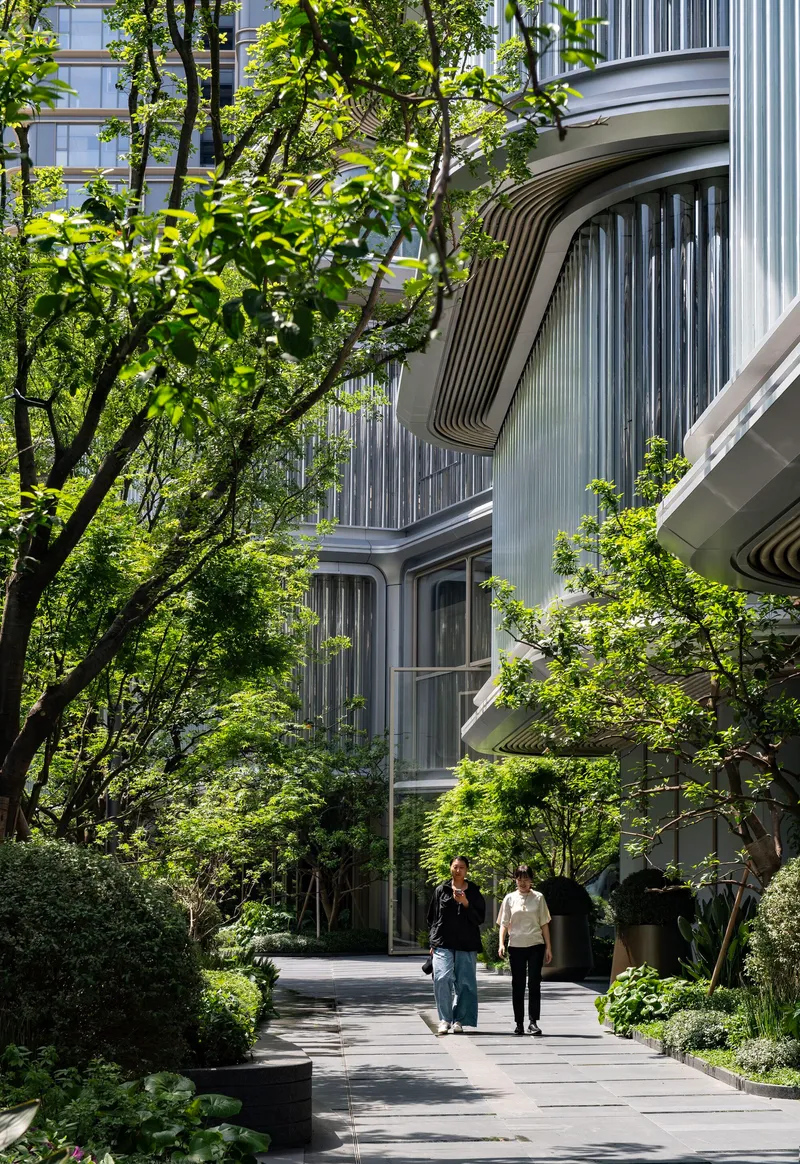Curved balconies with vertical metal fins framed by mature trees and a landscaped pathway with pedestrians