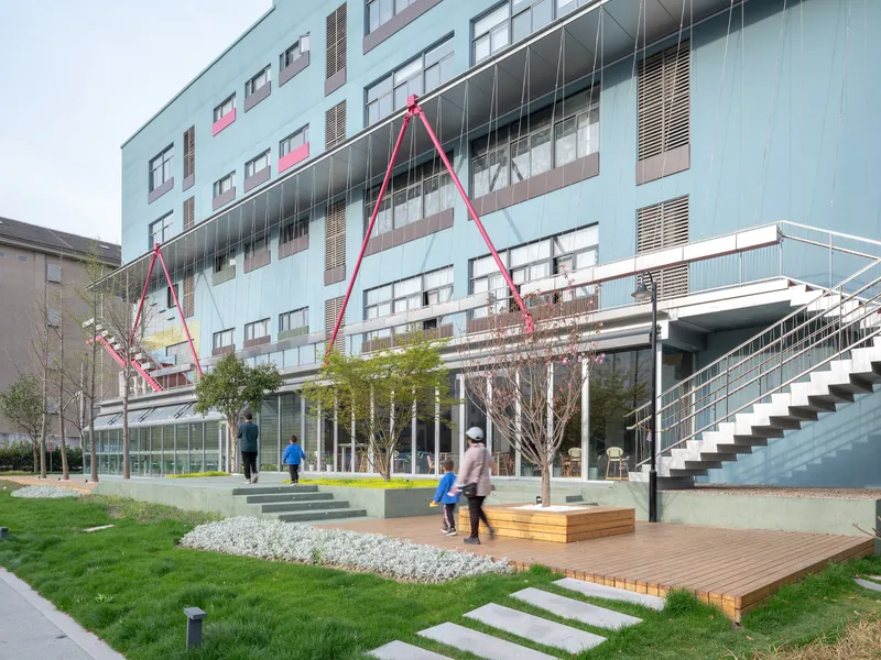 Ground-level courtyard with timber deck, planted lawn, and pink steel canopy structures with visitors passing