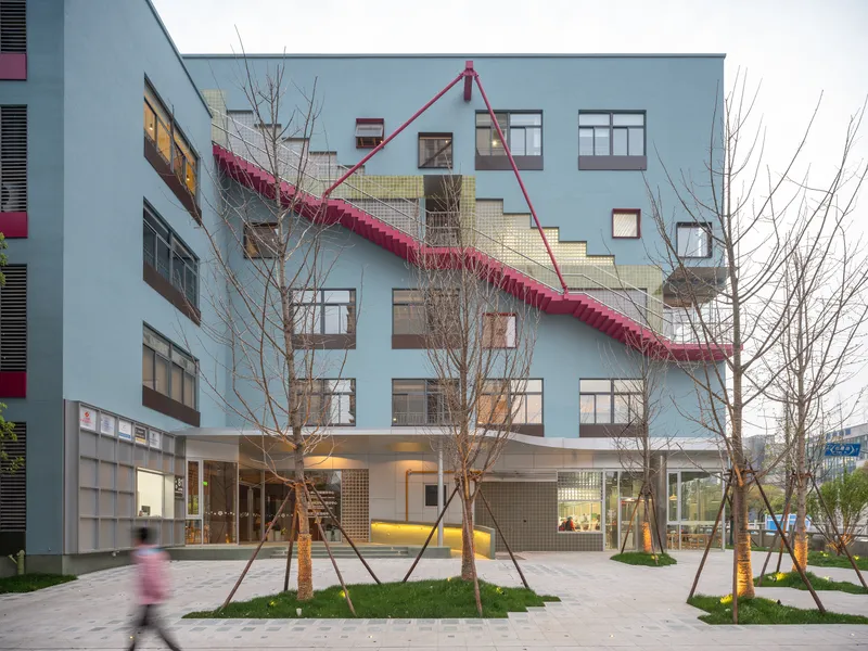 Street view of the pale blue facade with diagonal pink staircase and newly planted trees at dusk