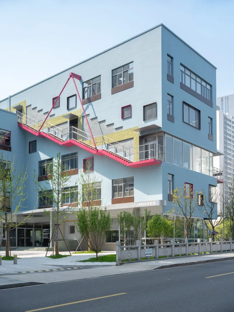 Corner facade showing the pale blue cladding, yellow tile band, and pink steel staircase with young street trees