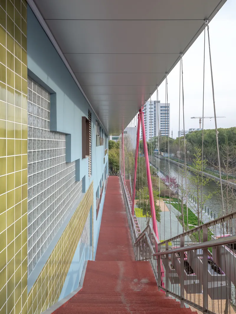 Suspended corridor with red resin flooring and cable railings running along yellow and blue tiled facade
