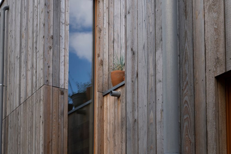 Weathered vertical timber cladding with narrow window opening and terracotta pot on exterior shelf under blue sky