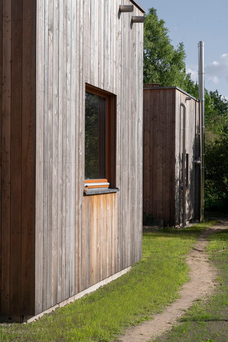 Weathered vertical timber cladding with window opening and timber shelf against cylindrical silo structure in grass clearing