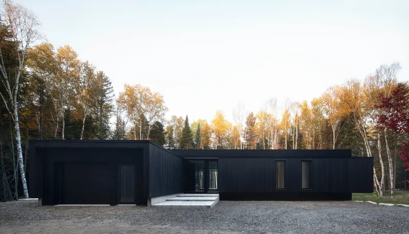 Front facade with vertical black cladding and stone entry steps set on a gravel forecourt among deciduous trees