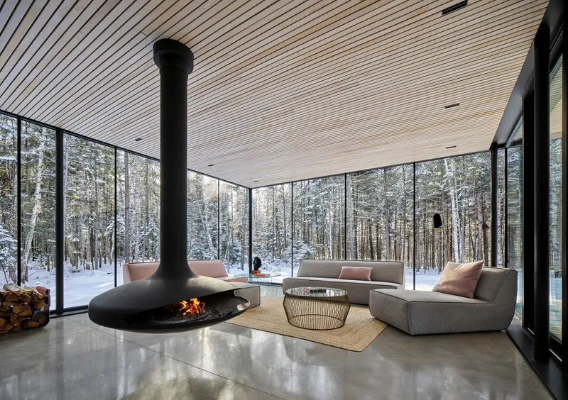 Living area with suspended conical fireplace and floor-to-ceiling glazing overlooking snow-covered forest