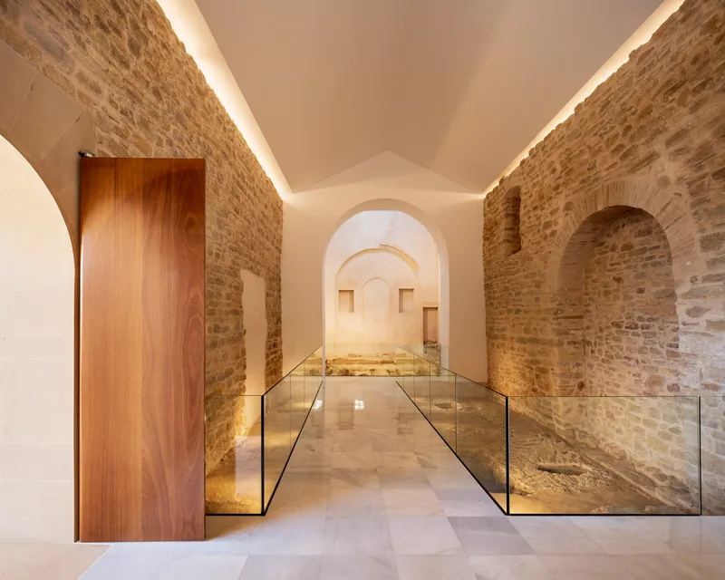 Vaulted corridor with exposed stone walls, walnut door and glass-edged floor revealing archaeological remnants below