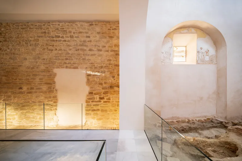 Arched niche in a white plaster wall with glass floor protecting excavated stone foundations beneath