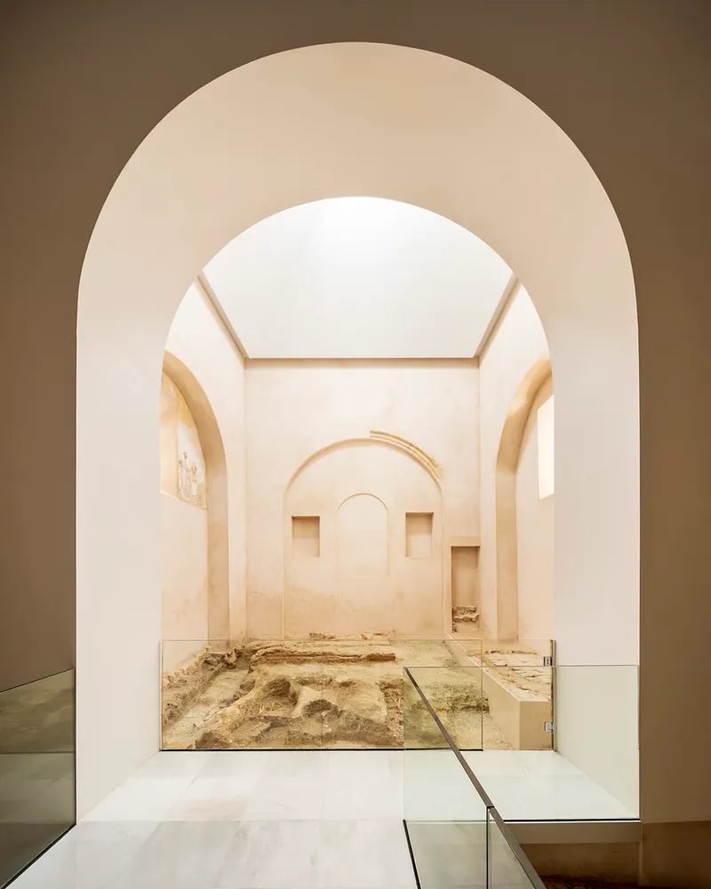 View through layered arches into a whitewashed room with glass-protected archaeological floor and flanking niches