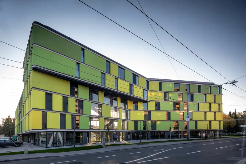 Long facade showing the chequered yellow and green panel composition with ground-level pilotis beneath a cloudy sky