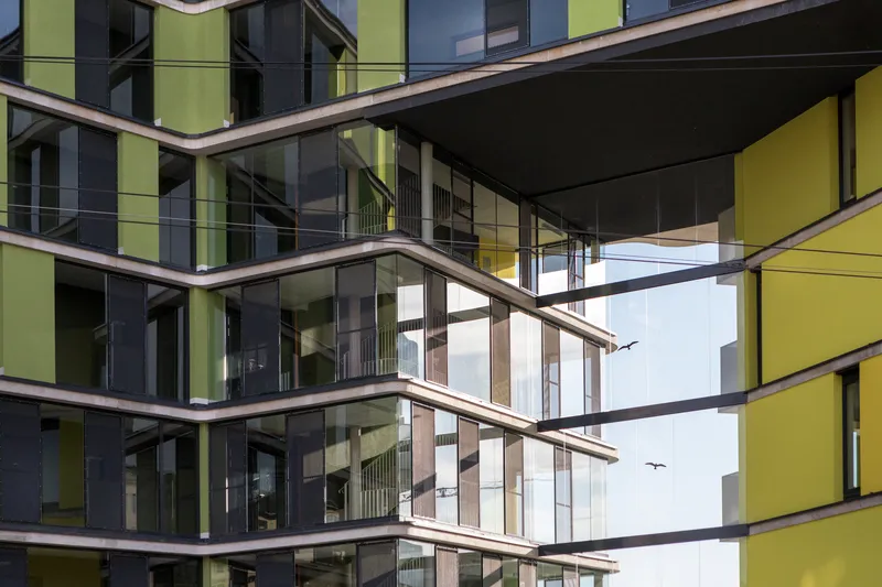 Corner facade with glass balconies framed by yellow and grey panels with birds flying overhead