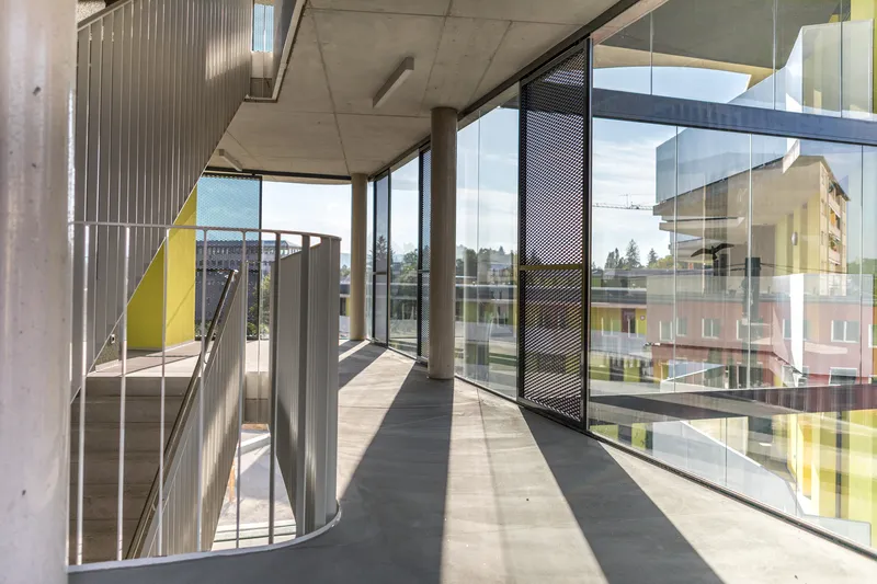 Interior corridor with exposed concrete ceiling, perforated metal screens and floor-to-ceiling glazing casting sunlight