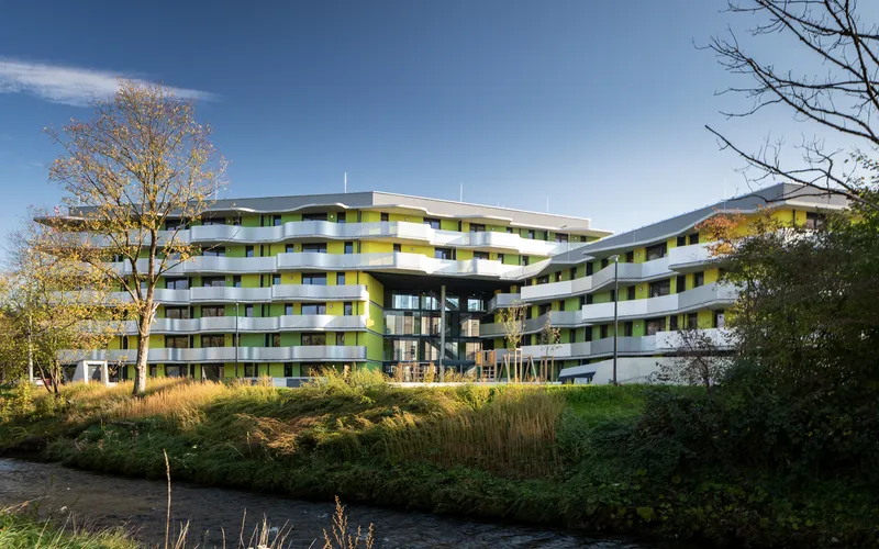Horizontal residential complex with yellow and green balcony panels beside a stream and bare trees