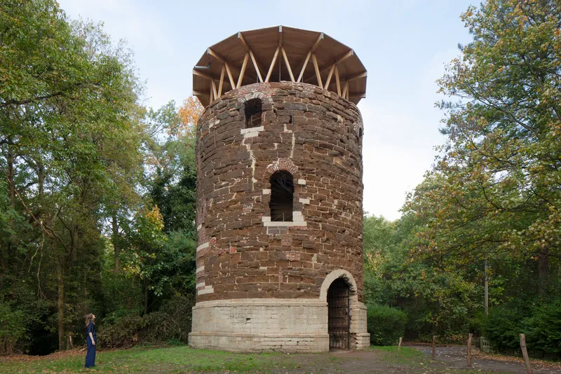 Cylindrical brick tower with zigzag timber roof structure framed by trees on a grassy clearing