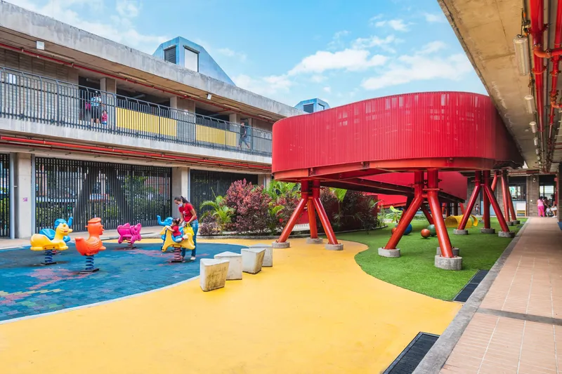 Children playing in a courtyard with blue wading pool and yellow rubber surfacing beneath the curved red pedestrian bridge