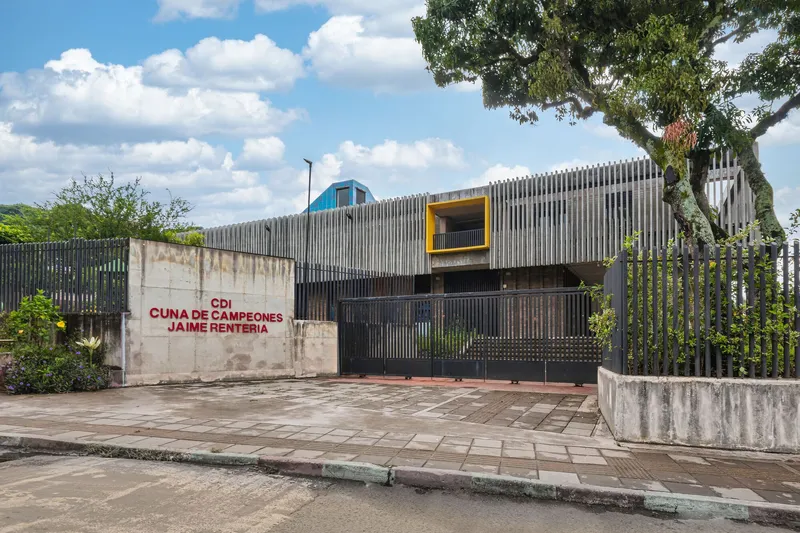 Street view of vertical metal slat facade with yellow window frame and blue pyramid roof beside weathered concrete wall