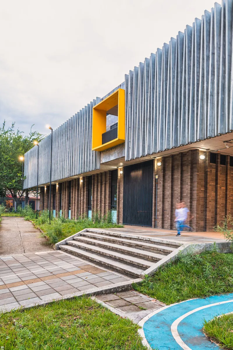 Entrance facade with vertical corrugated metal cladding, yellow window aperture, and brick base at dusk
