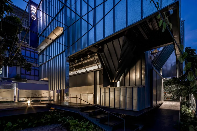 Corner pavilion with folded metal roof and vertical cladding lit from within at twilight