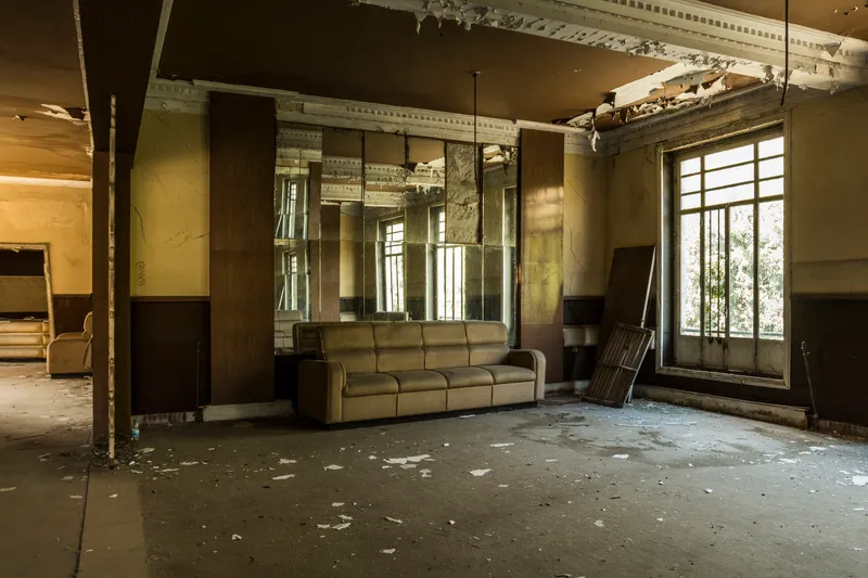 Abandoned interior hall with ornate ceiling moldings, tall windows, and a worn sofa under natural light