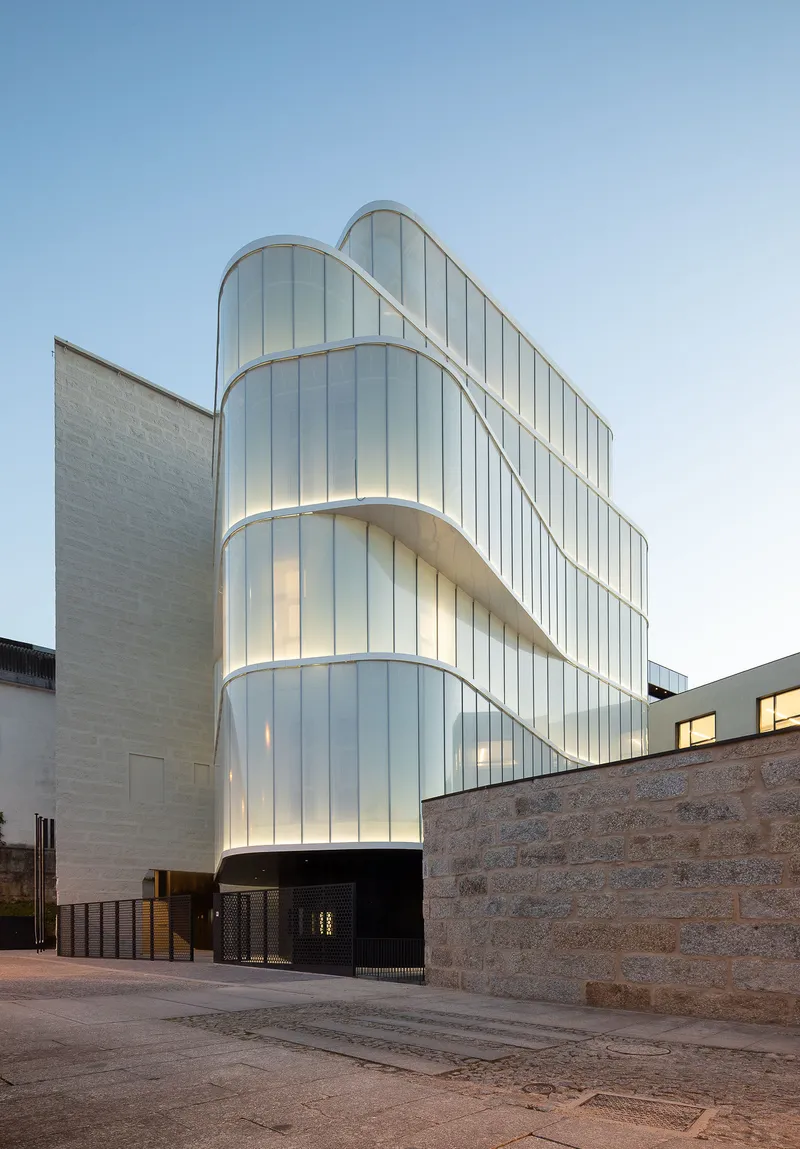 Curved translucent glass tower with vertical fins rising above stone walls at dusk