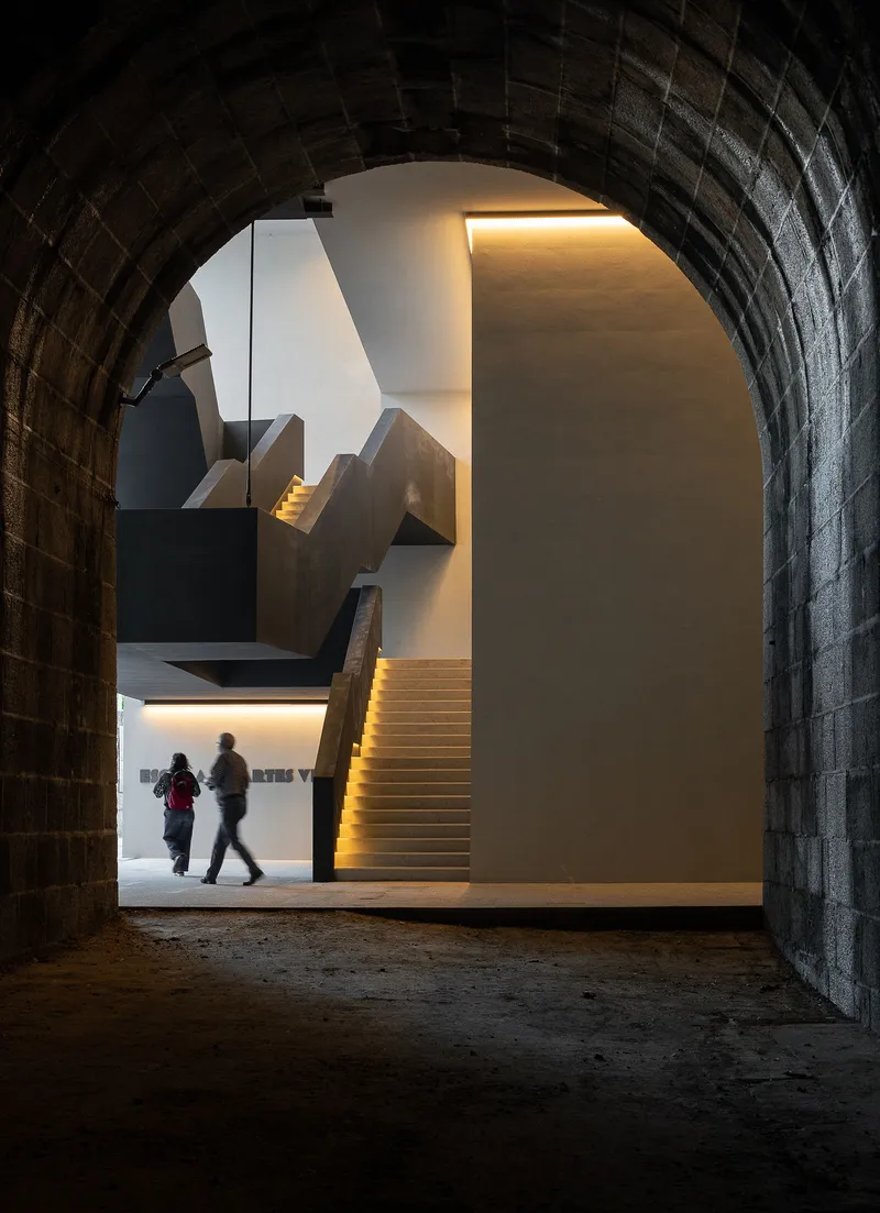 View through barrel-vaulted granite passage toward illuminated staircase with two visitors walking