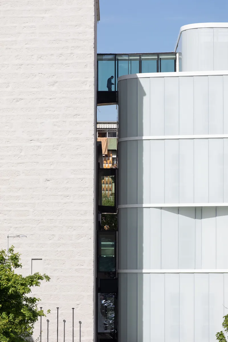 Narrow gap between white brick wall and cylindrical white-paneled facade revealing person on upper balcony