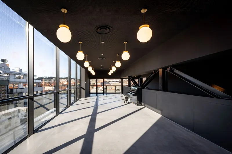 Upper floor corridor with dark perforated ceiling, globe pendant lights, and glazed facade overlooking rooftops