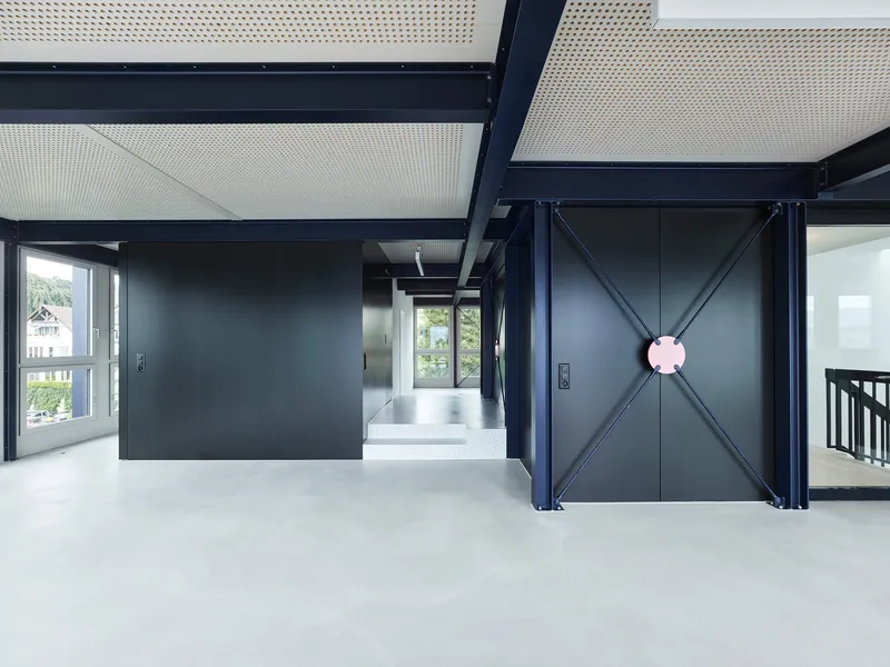 Open plan interior with exposed steel frame, perforated ceiling panels, and black partition walls with gray flooring