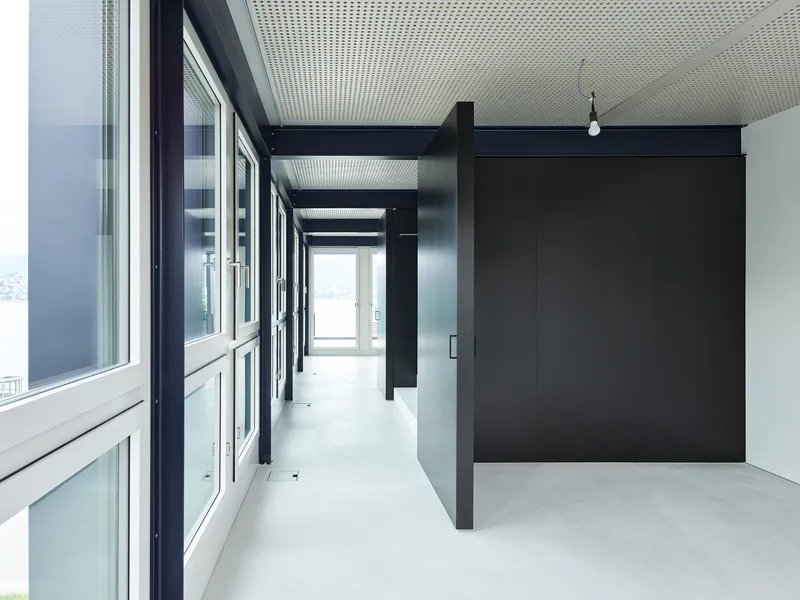 Circulation corridor with black steel columns, glazed wall along one side, and perforated ceiling panels filtering daylight