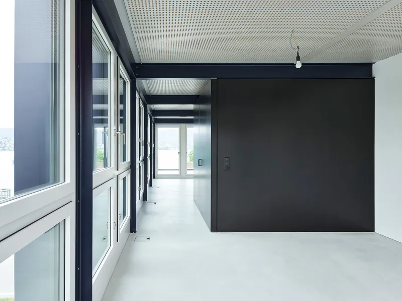 Hallway view along the glazed facade with a dark partition volume and perforated ceiling