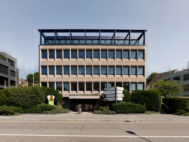 Street view of the layered facade with terracotta panels and a recessed glazed penthouse above green hedges