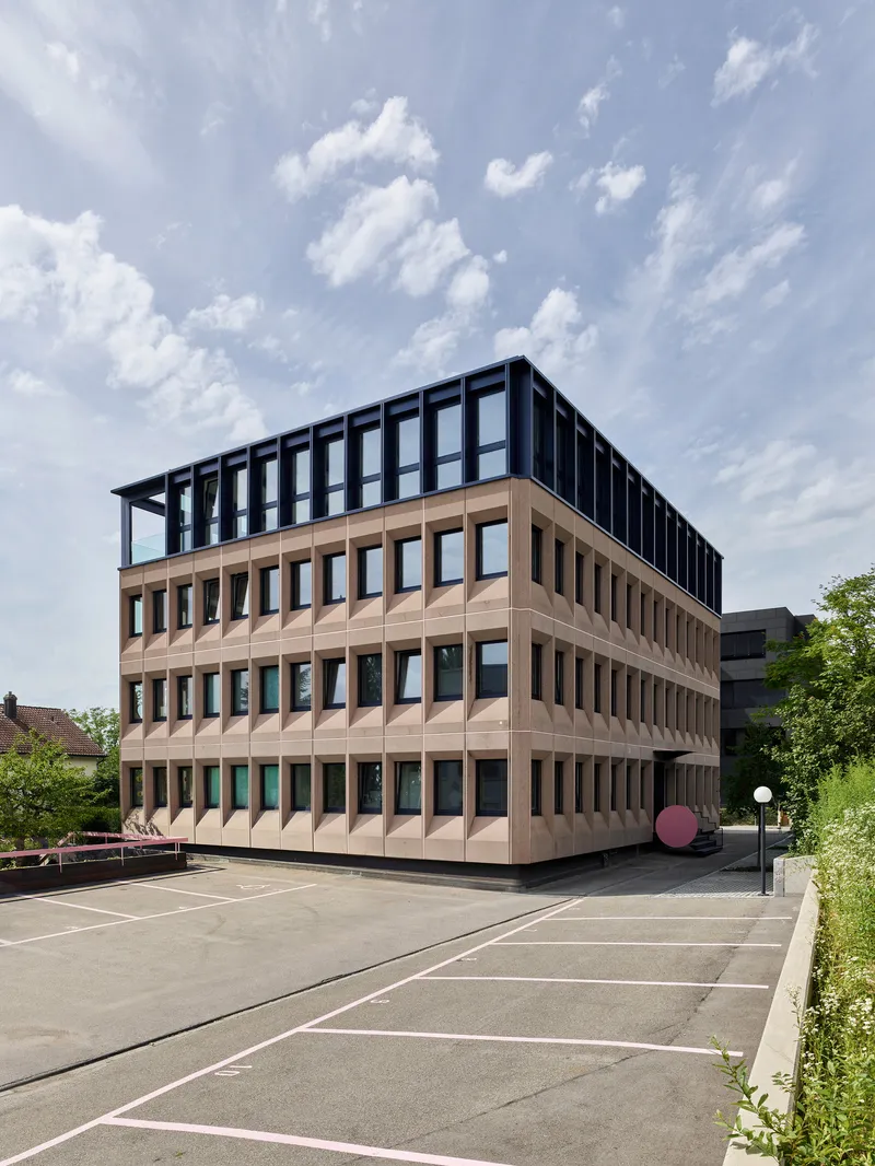 View across the parking area showing the folded terracotta facade and rooftop steel frame under blue skies