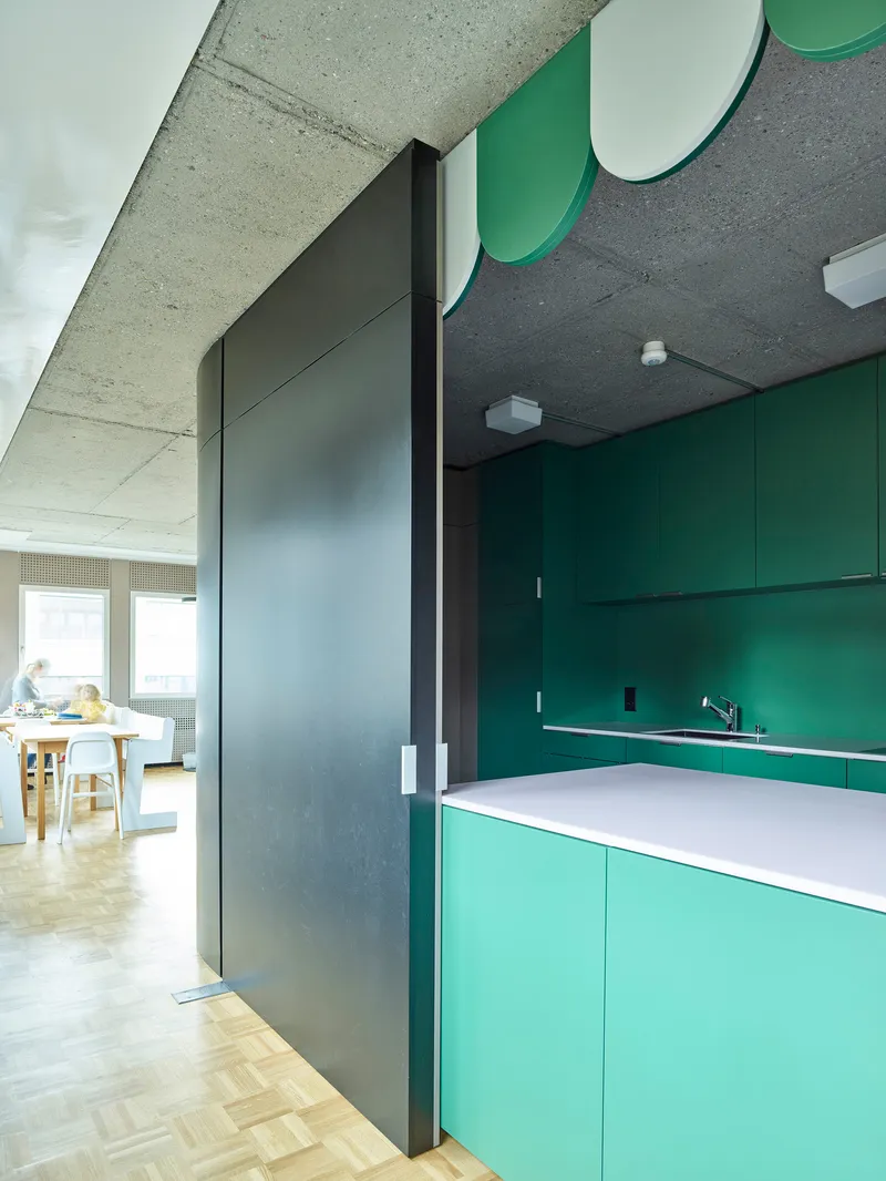Kitchenette with green and mint cabinets beneath exposed concrete soffit with scalloped green detail