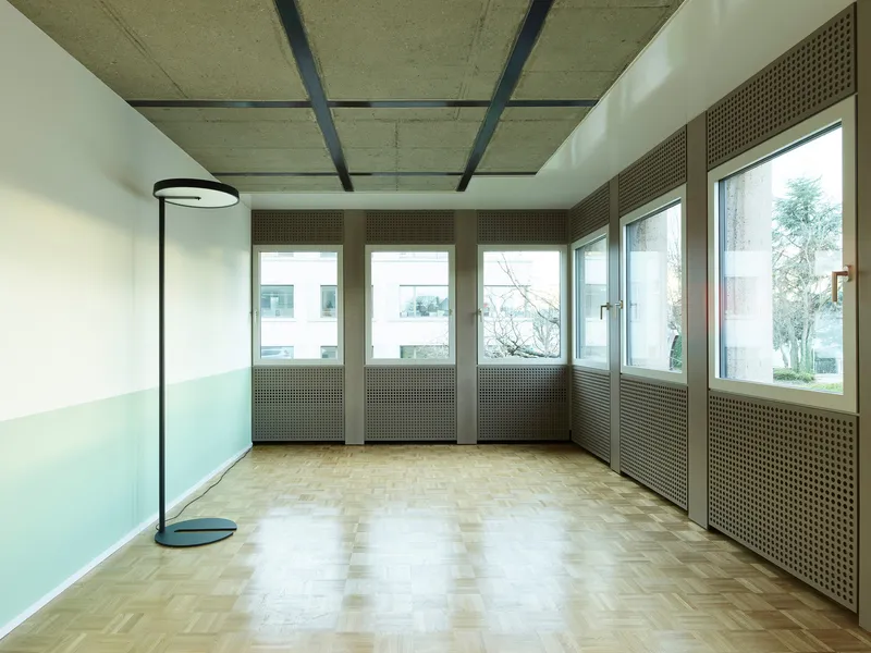 Room with perforated radiator covers below windows, parquet floor, and concrete ceiling with steel beams