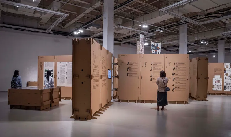 Visitor approaching cardboard partition walls with printed text as others explore the exhibition space