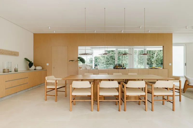 Dining room with long timber table and wood-paneled wall framing a horizontal window to greenery