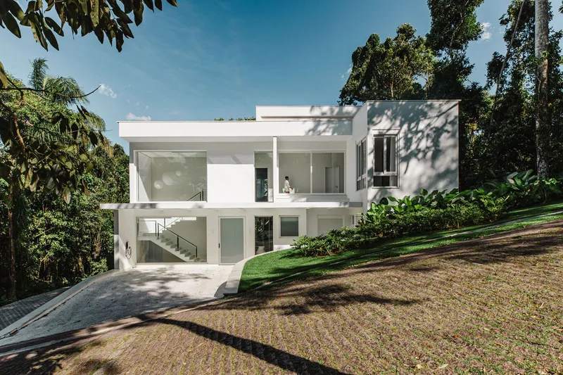 Two-story white volume set into a sloping lawn surrounded by tall trees under afternoon sun