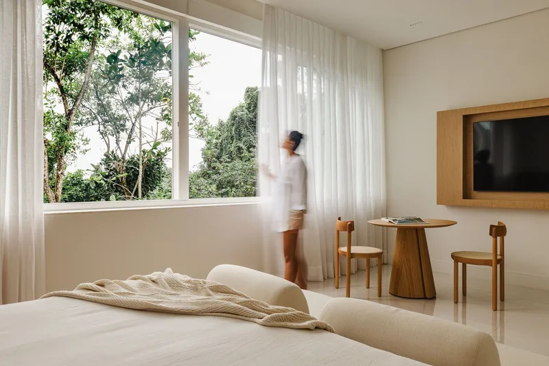 Bedroom with a figure in motion near the corner window overlooking dense green foliage