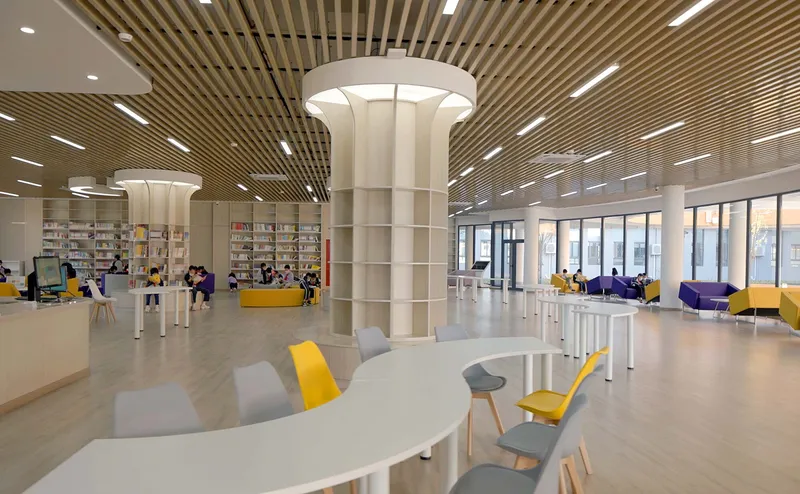 Library interior with slatted timber ceiling, white columns and scattered seating with yellow and grey chairs