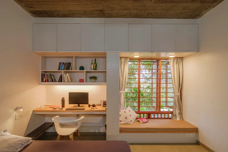 Bedroom workspace with built-in desk, white storage wall and window seat overlooking green foliage through latticed timber shutters