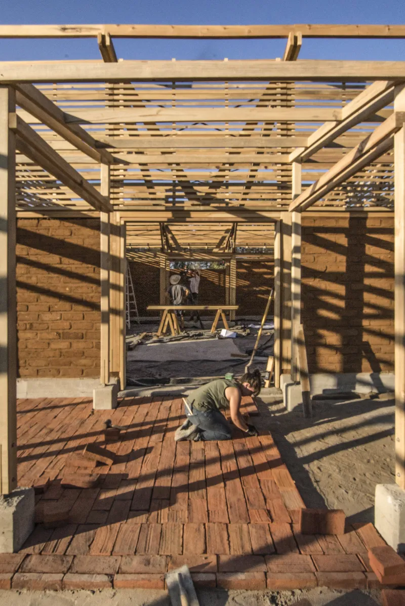 Worker laying brick flooring within the timber frame structure casting geometric shadow patterns at dusk