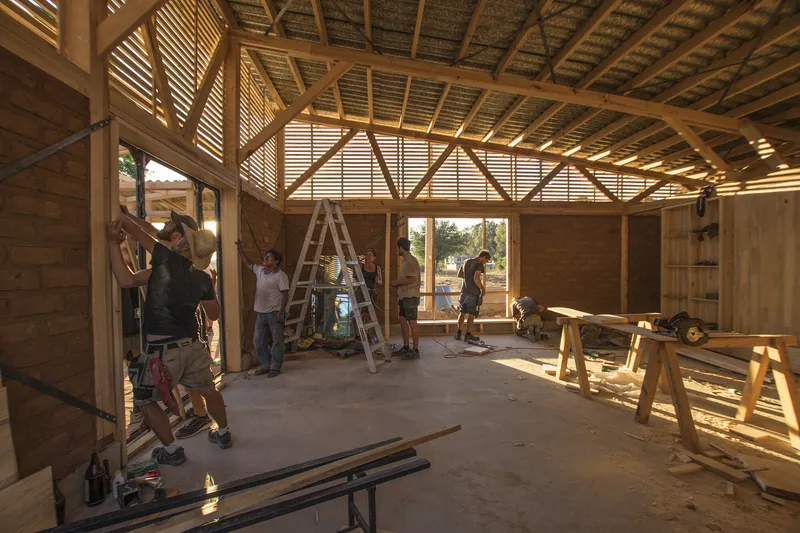 Interior space under construction with exposed timber trusses and workers installing wall panels in afternoon light