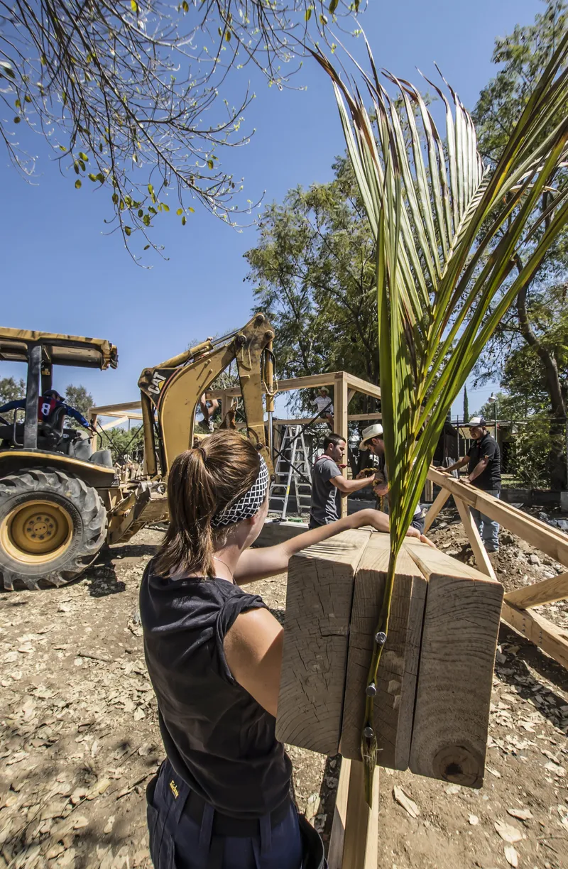 Construction site with workers assembling timber framework and palm fronds under a clear blue sky