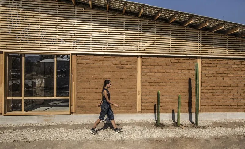 Rammed earth wall facade with horizontal timber louvers above and a woman walking past cacti