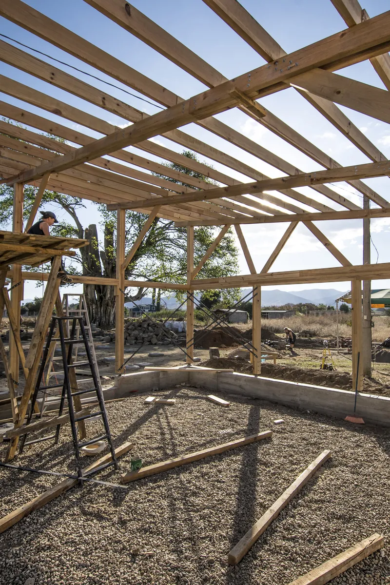Exposed timber frame structure under construction with diagonal bracing and workers on ladders beneath open sky