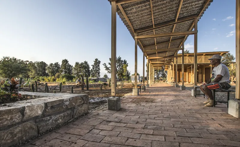 Covered walkway with timber columns and slatted roof over terracotta brick pavers with workers resting nearby