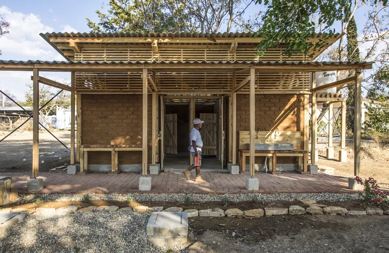 Front facade of a timber-frame pavilion with corrugated metal roof and a person standing in the doorway