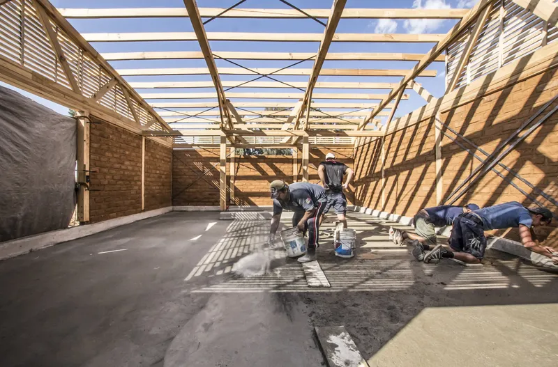 Interior space with workers applying concrete floor finish under diagonal shadows from timber roof trusses above