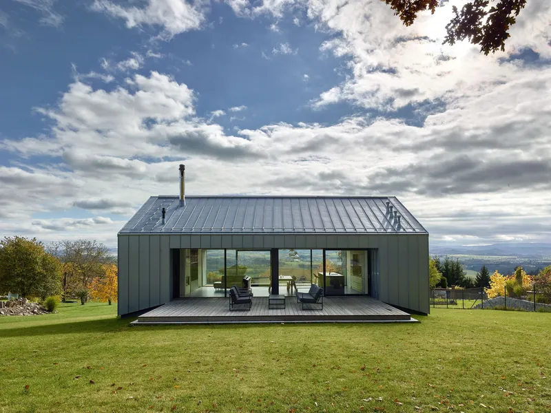 Metal-clad gabled volume with timber deck and full-height glazing opening to a lawn under cloudy skies