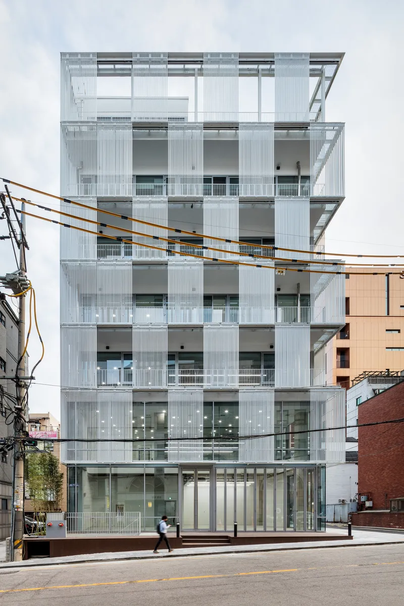 Front elevation showing the layered balconies behind vertical corrugated screening with a person walking past the entrance